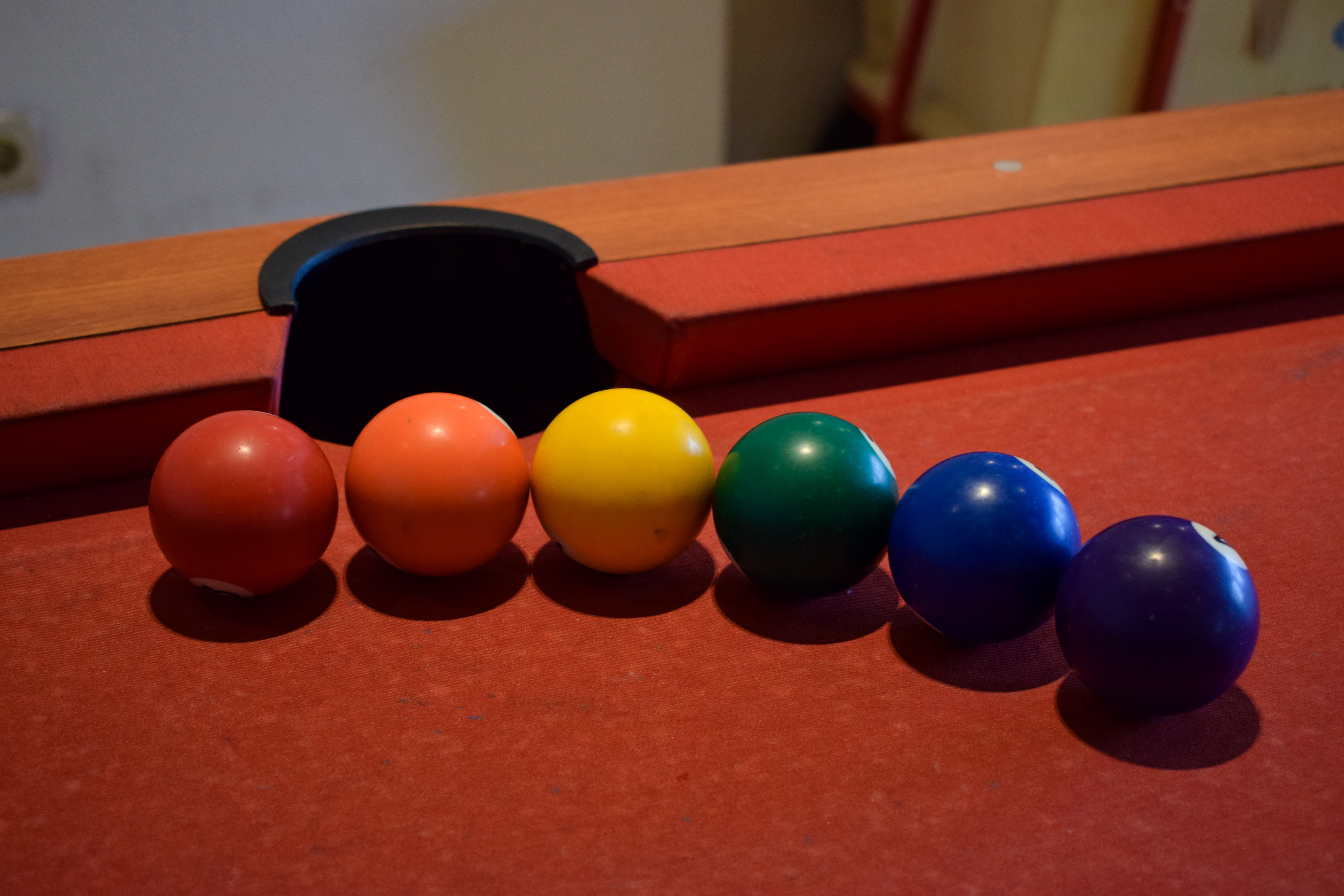 Es sind sechs Billardkugeln auf dem Billardtisch zu sehen. Sie sind von links nach rechts: rot, orange, gelb, grün, blau, lila