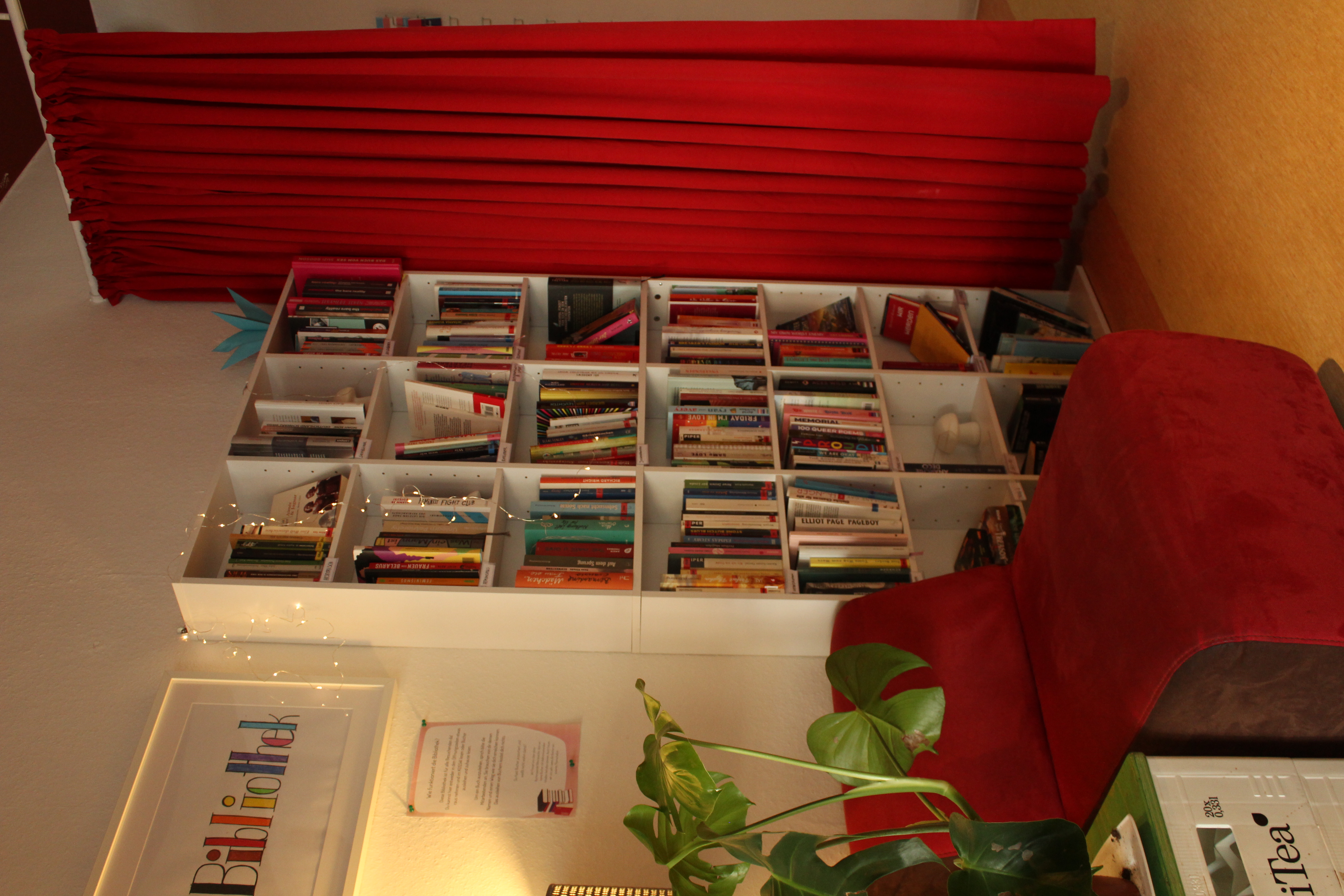 Im Zentrum des Bildes ist ein Bücherschrank und ein Sessel zu sehen. Über dem Sessel hängt ein Bilderramen in dem Bibliothek steht. rechts neben dem Regal hängt ein roter Vorhang